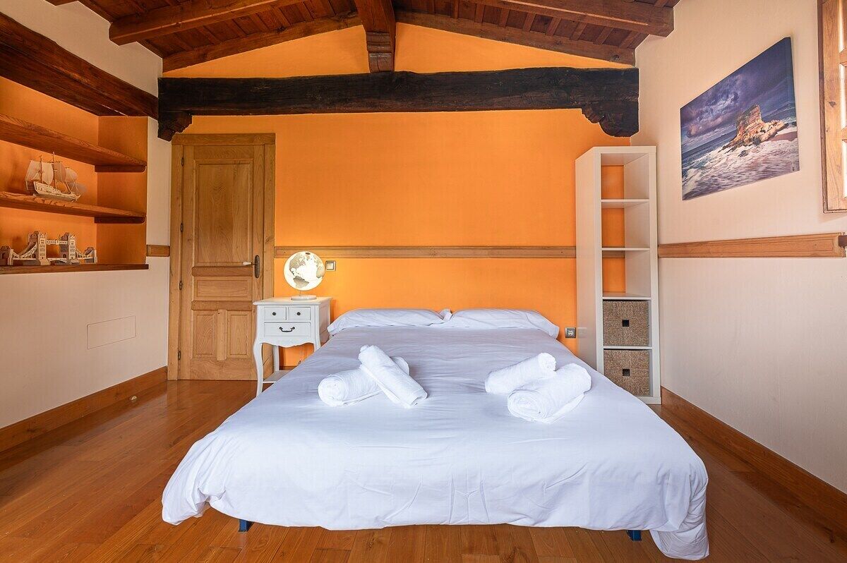 Una cama bien hecha con sábanas y toallas blancas, una puerta de madera, una mesita de noche con una lámpara y una estantería con objetos decorativos.