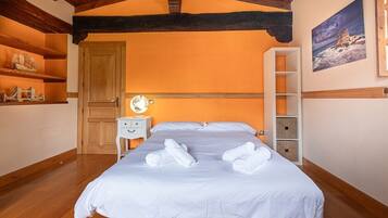 Una cama bien hecha con sábanas y toallas blancas, una puerta de madera, una mesita de noche con una lámpara y una estantería con objetos decorativos.