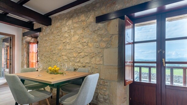 Un comedor con pared de piedra, vigas de madera en el techo y vista a través de un amplio ventanal.