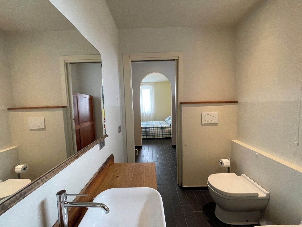 Un baño con lavamanos, espejo e inodoro. Un pasillo conduce a un dormitorio con cama y ventana.