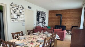 Een eethoek met een tafel voor vier personen, omringd door houten stoelen. Op de achtergrond staat een bank met een rode kussen en een groen sierkussen. Boven de bank hangt een groot zwart-wit abstract schilderij.