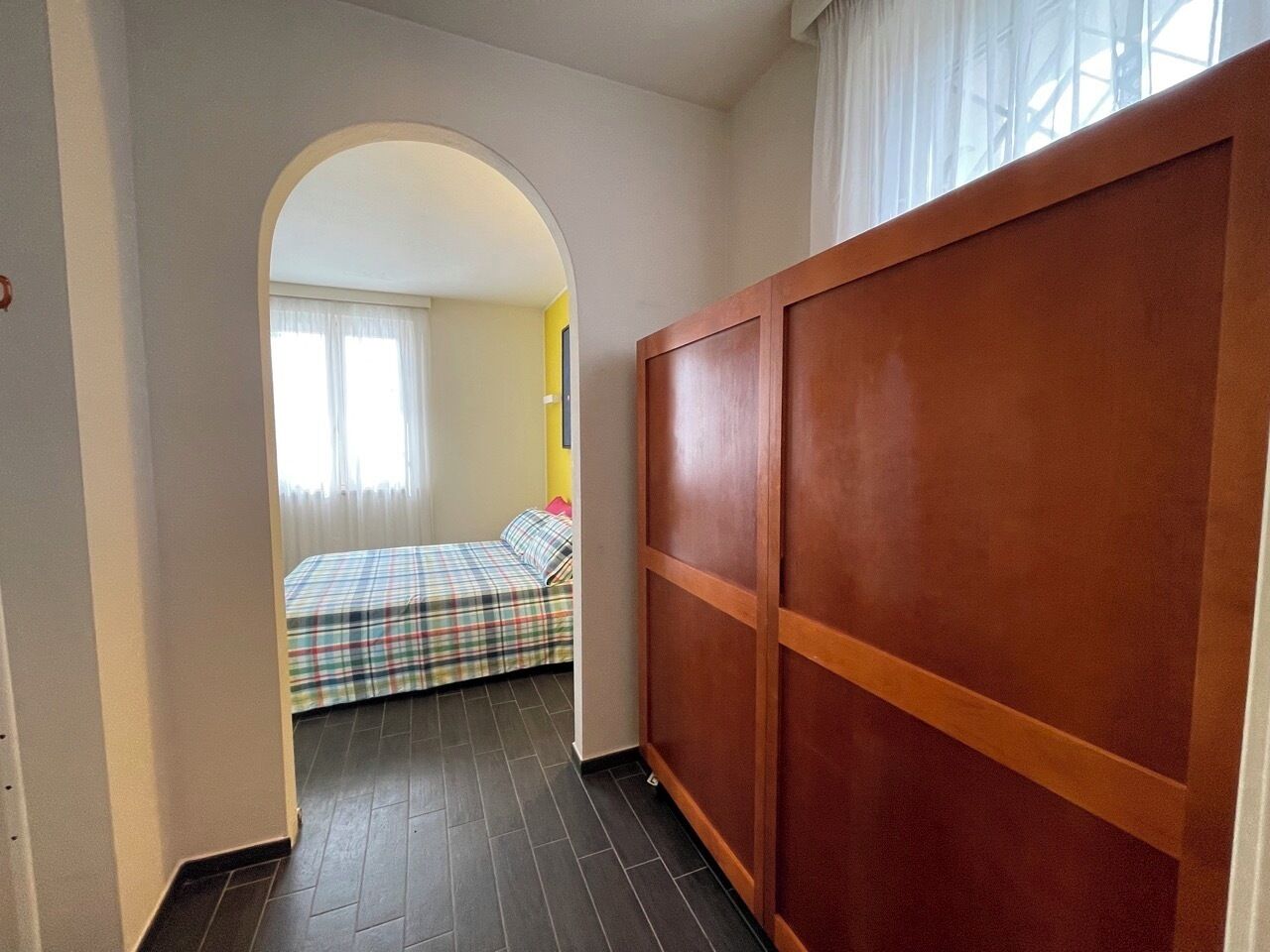 A narrow hallway leading to a bedroom with a bed, a wooden wardrobe, and a window with curtains.