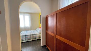A narrow hallway leading to a bedroom with a bed, a wooden wardrobe, and a window with curtains.