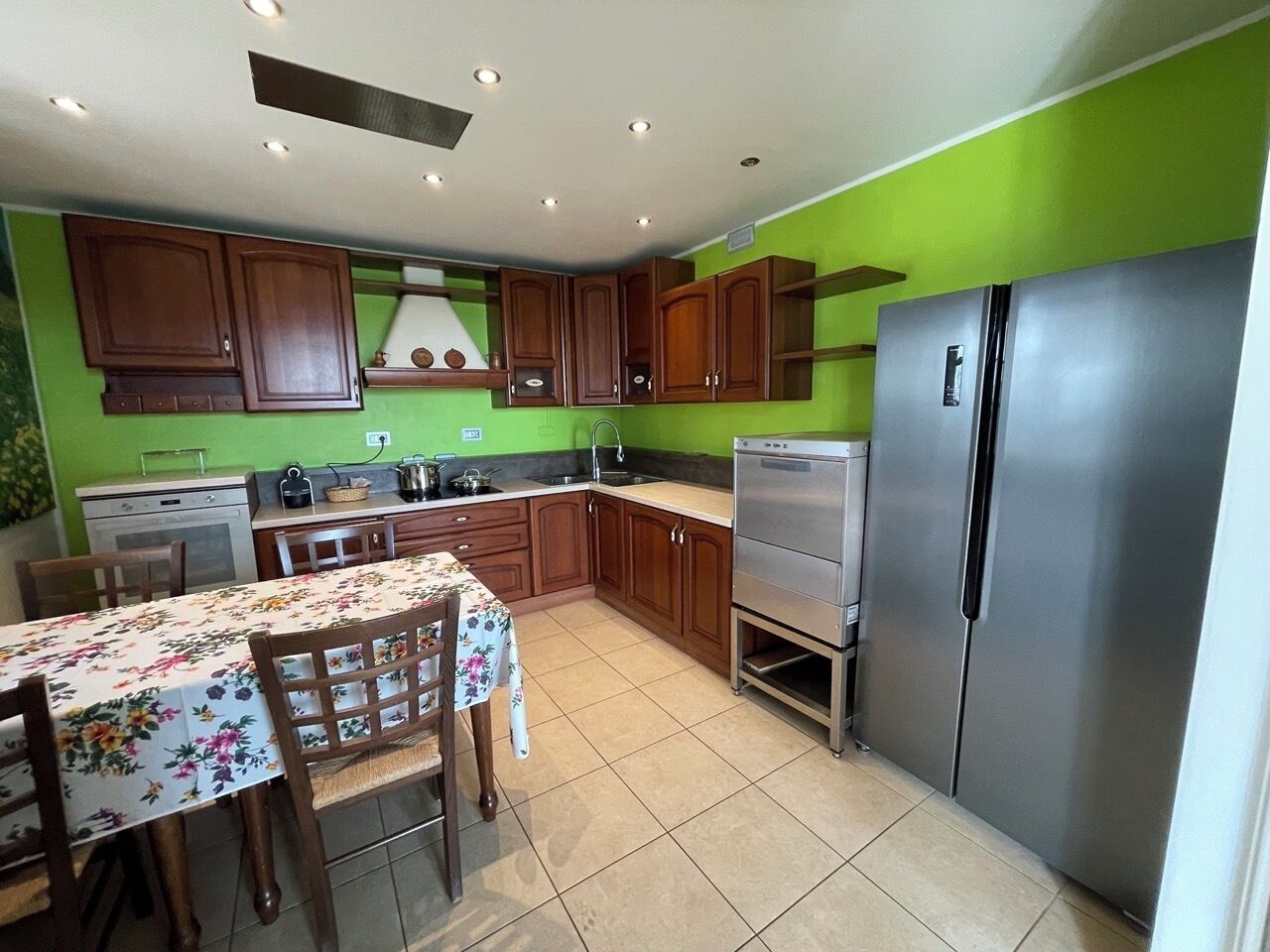 A kitchen with wooden cabinets, a stainless steel refrigerator, a dining table with chairs, and a tiled floor.