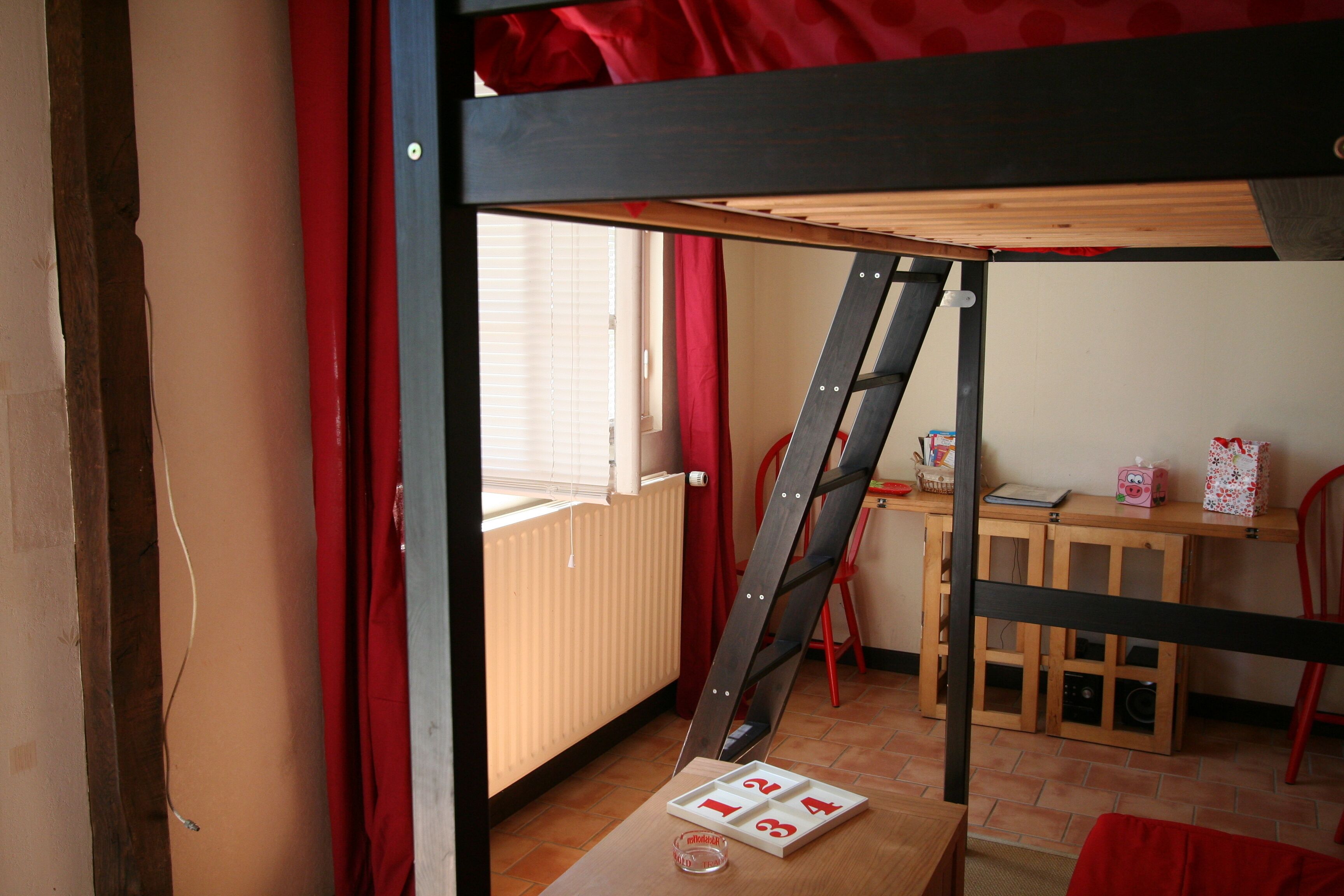 Une chambre avec un lit superposé, une échelle en bois, une petite table avec un jeu de cartes, une chaise et une fenêtre avec des stores.
