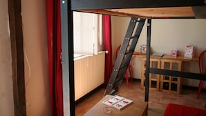Ein Schlafraum mit Etagenbetten, einer Holzleiter, einem kleinen Tisch mit einem Kartenspiel, einem Stuhl und einem Fenster mit Jalousien.