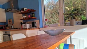 Una cuina moderna amb taula de fusta, cadires blanques i vista a arbres a través de grans finestres.