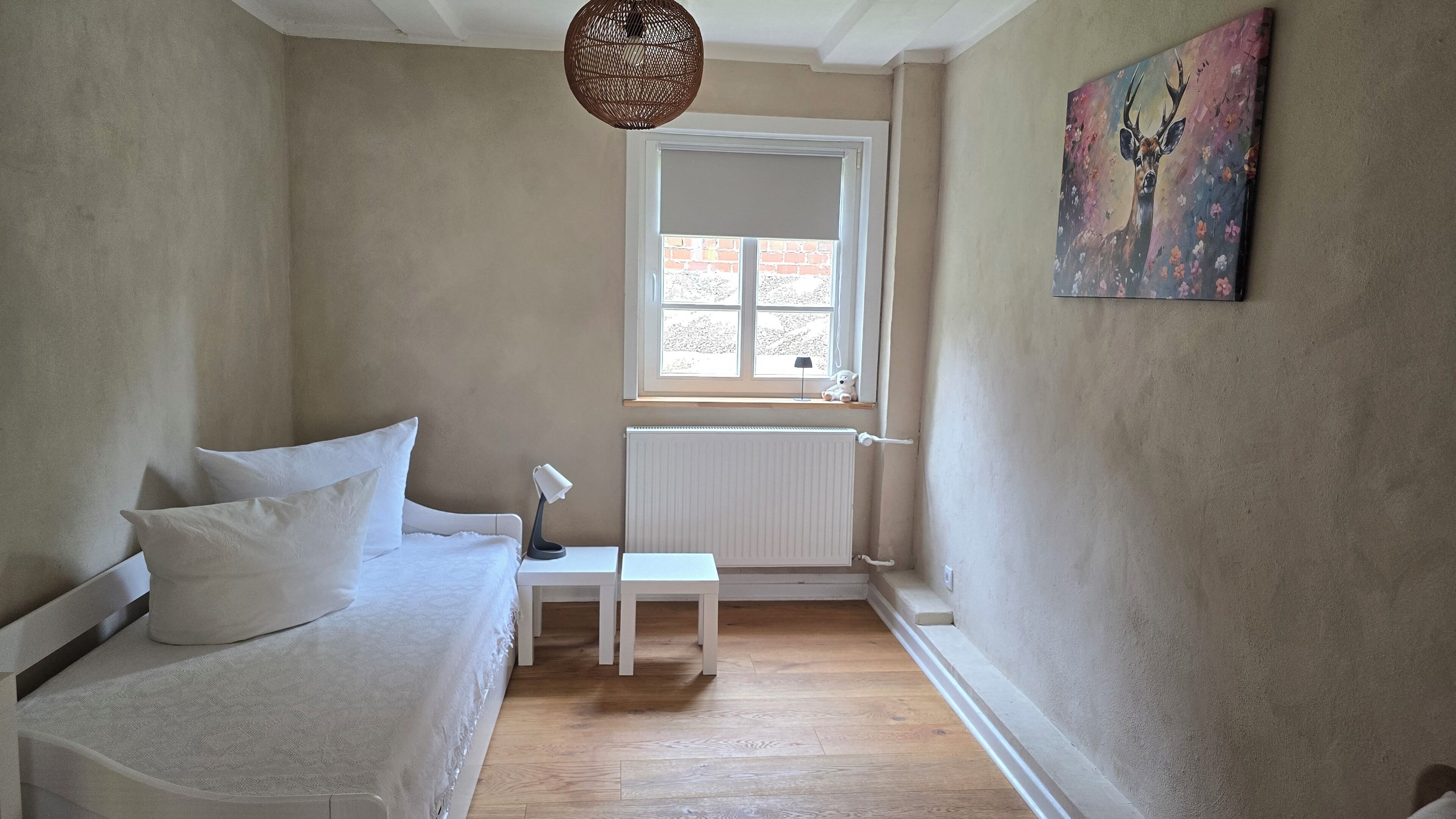 Quarto com cama, mesa de cabeceira, luminária, radiador, janela e um quadro na parede.