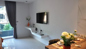 A modern living room with a flat-screen TV mounted on the wall, a white console table with decorative items, a dining table with a vase of flowers, and a view of the outdoors through a large window. - Quiet condo with pool vibe (Tambon Patong)