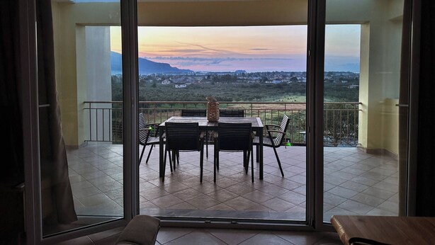 Een balkon met een tafel en stoelen, een uitzicht op een landschap met huizen en bergen, en een heldere lucht.