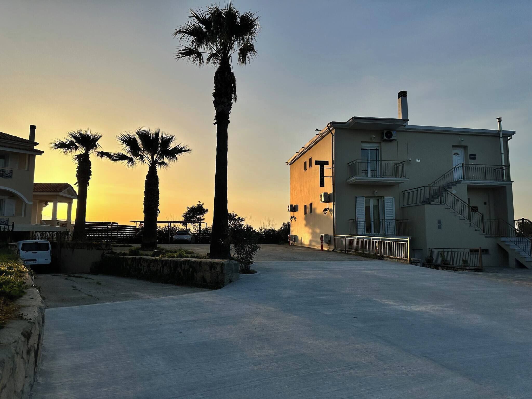 A building with a balcony, a staircase, and a parking area with palm trees.