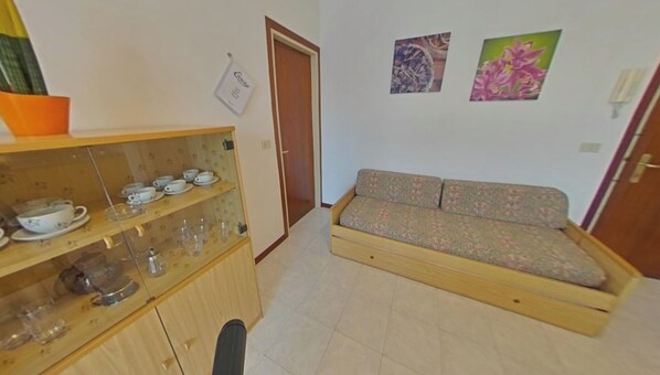 A compact living space with a sofa, a cabinet with dishes, and two wall-mounted pictures. - Convenient flat in a great location (Lignano Sabbiadoro)