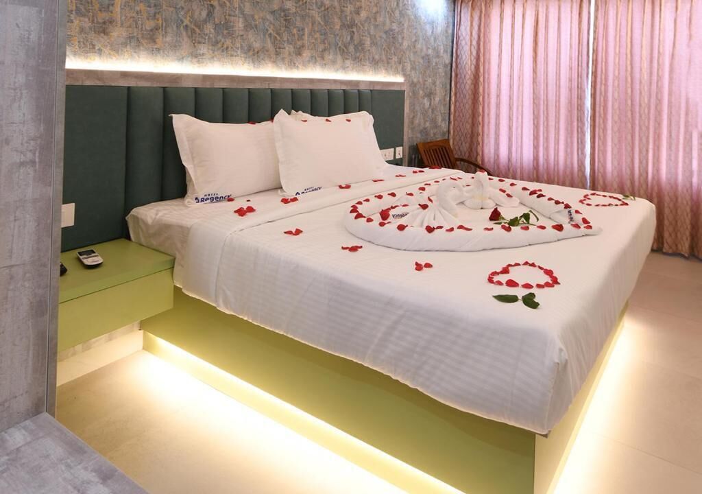 Un lit bien fait, décoré de pétales de roses et agrémenté d’une composition florale en forme de cœur, une table de chevet verte avec un téléphone, et une tête de lit adossée à un mur texturé.