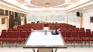 A conference room with rows of red chairs, a central table with a microphone, and a speaker mounted on the wall. - Hotel Regency Grande (Thiruvananthapuram)