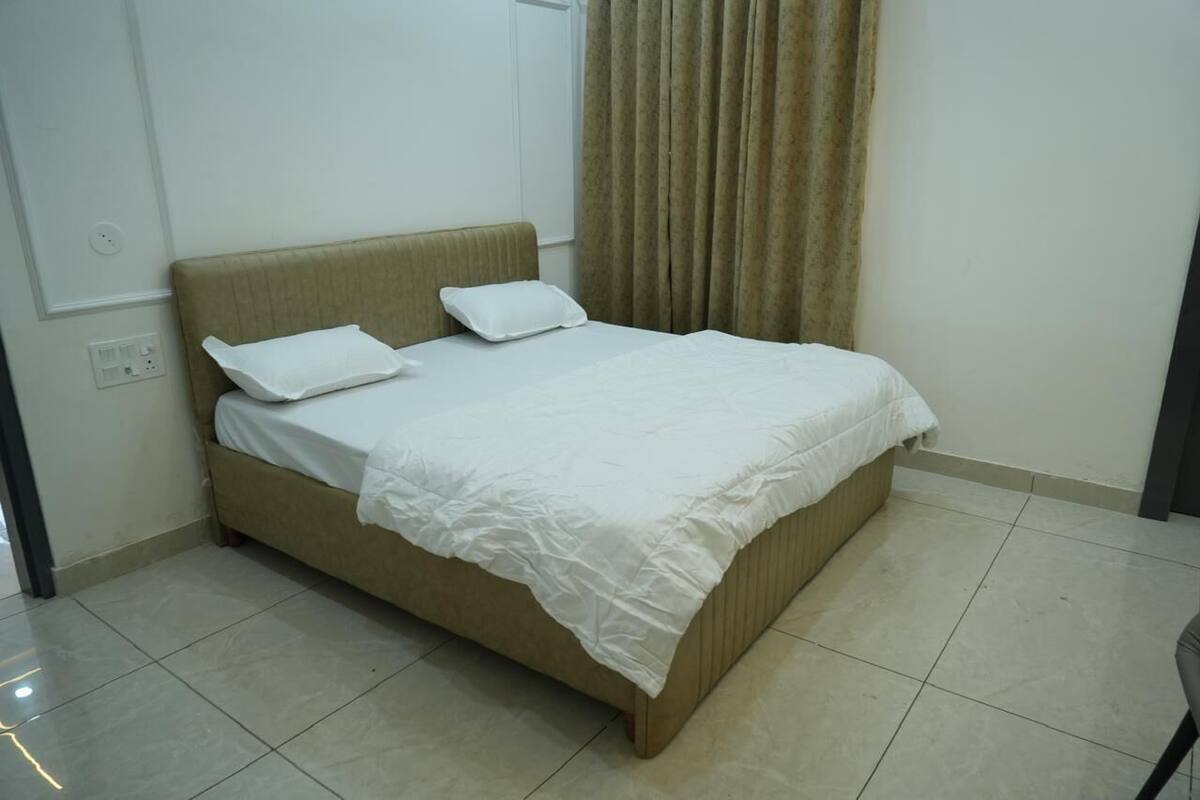 Un lit simple avec une couvre-lit blanche et deux oreillers, une tête de lit beige et un rideau à une fenêtre.