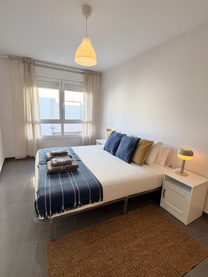 A modern bedroom with a large bed, a nightstand with a lamp, a window with curtains, and a view of the outside. - Dragos Suites 104 (Arrecife)