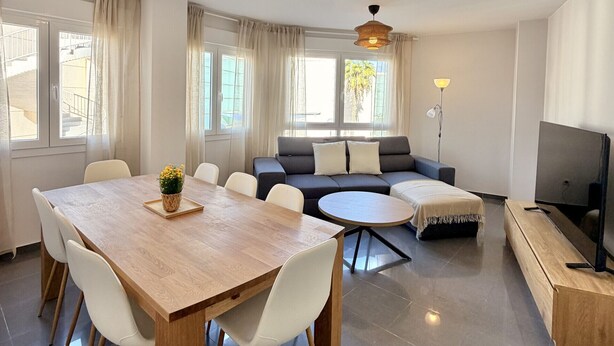 Uma sala de estar moderna com mesa de jantar, sofá, televisão e grandes janelas.