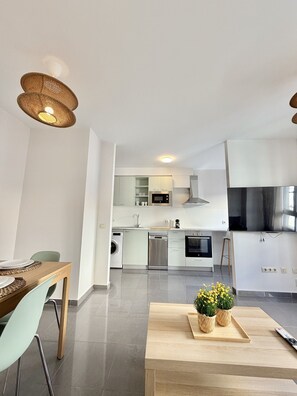 A modern kitchen with stainless steel appliances, a dining area with a wooden table and chairs, and a living space with a sofa and a coffee table. - Dragos Suites 103 (Arrecife)