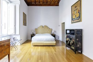 A bedroom with a wooden floor, a bed with a patterned headboard, bedside tables with lamps, a dresser, and a decorative cabinet with floral artwork. - Luxury Apartment in Piazza Farnese | Walk to Everything | Quiet & Central (Roma)