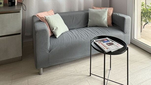 Un salon moderne avec un canapé gris, une petite table noire sur laquelle sont posés des magazines et une fenêtre ornée de rideaux légers.