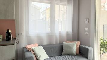 Ein modernes Wohnzimmer mit einem grauen Sofa, einem kleinen schwarzen Tisch mit Zeitschriften und einem Fenster mit transparenten Vorhängen.