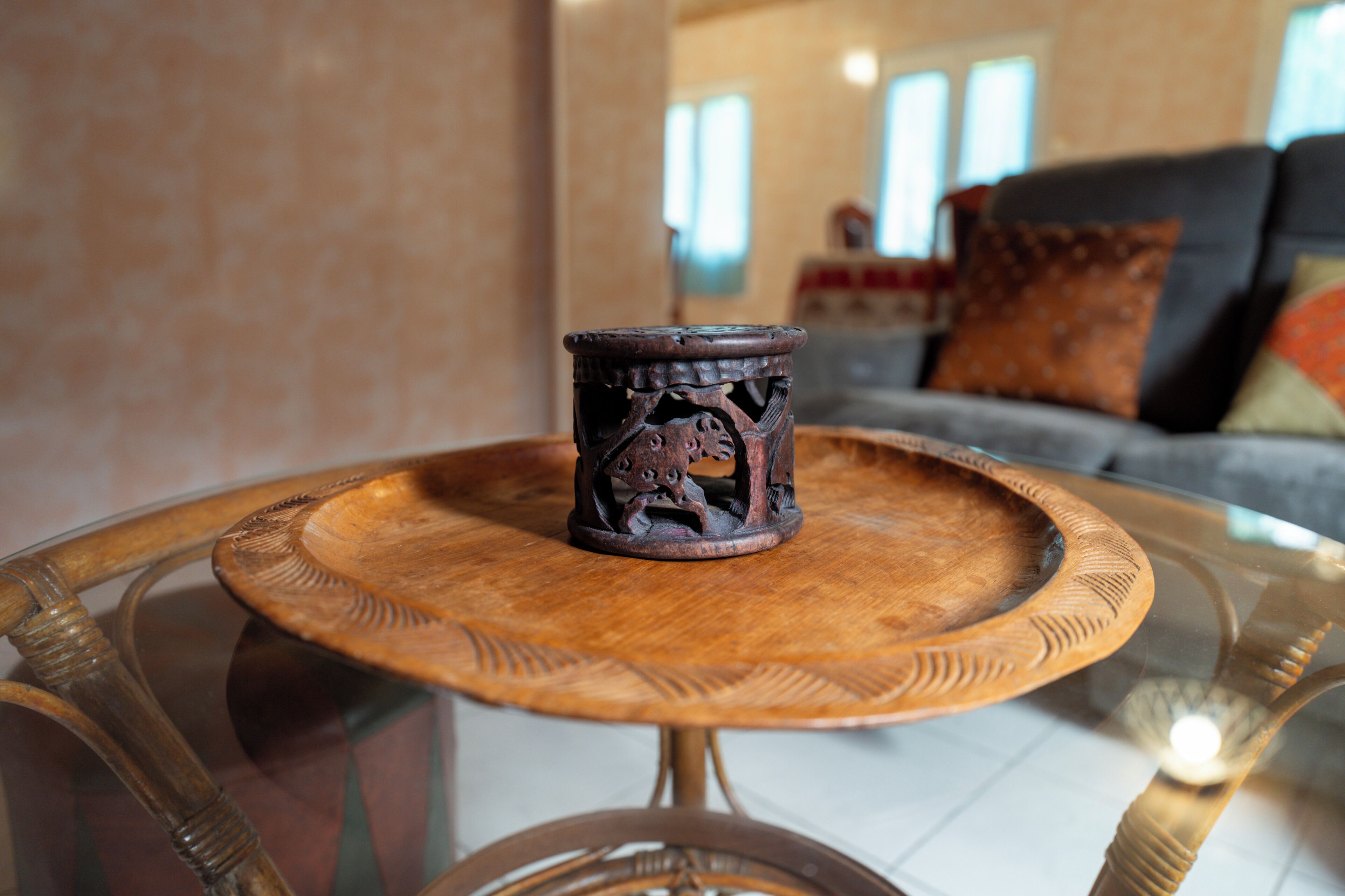 Una mesa redonda de madera con cubierta de vidrio, una taza de madera decorativa y un sofá con cojines al fondo.
