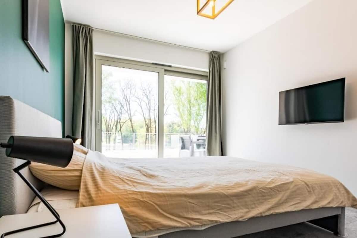 A modern hotel room with a bed, a desk, a lamp, a TV, and a large window.