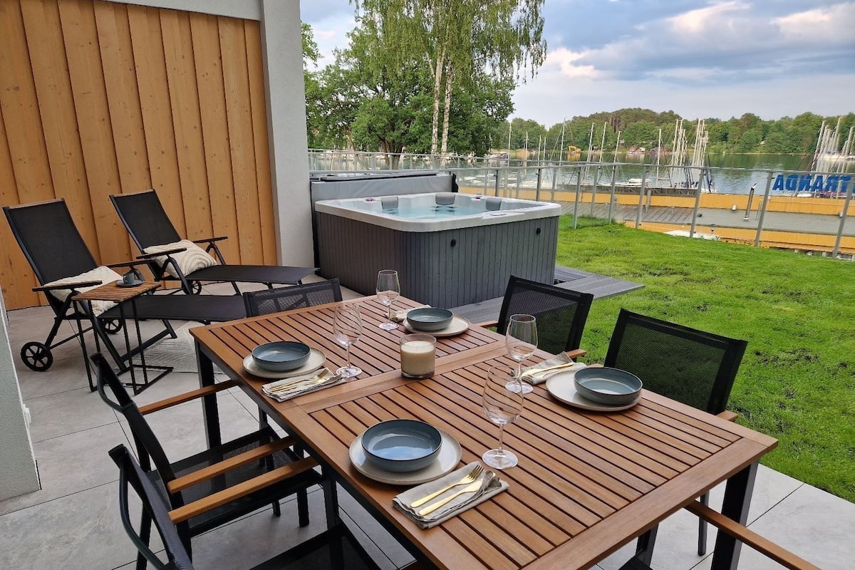 An outdoor dining area with a wooden table set for two, a hot tub, and lounge chairs.
