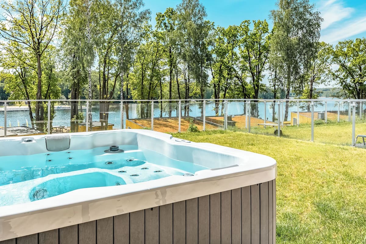 A hot tub with a wooden exterior, surrounded by a grassy area and a glass railing, overlooking a body of water and trees.