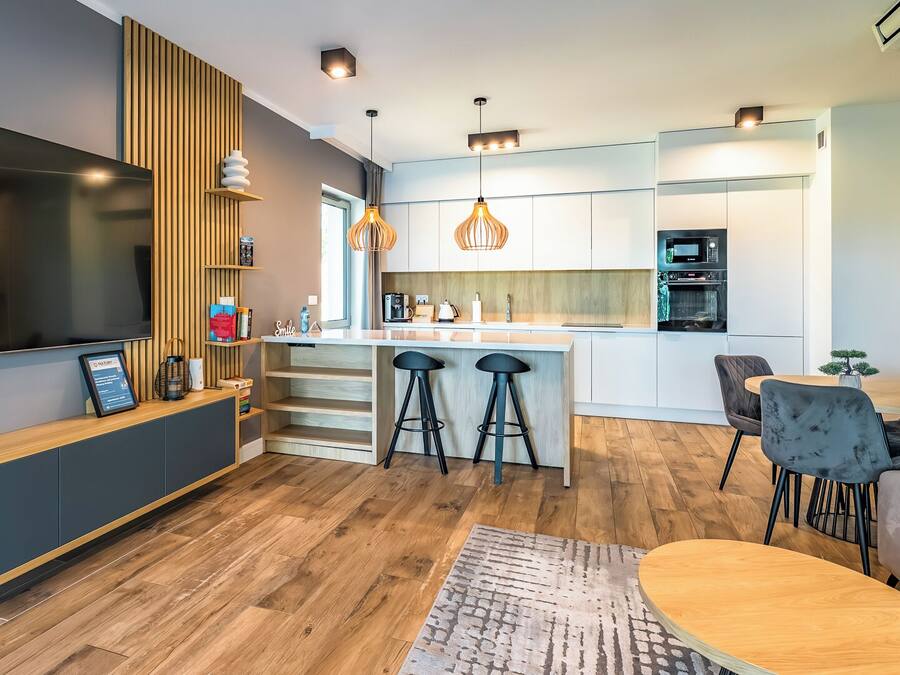 A modern kitchen with a central island, bar stools, a dining area with a table and chairs, and a TV mounted on the wall.