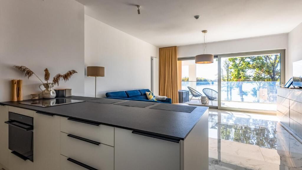 A modern kitchen with a central island, a sofa with cushions, and a large sliding glass door leading to a balcony.