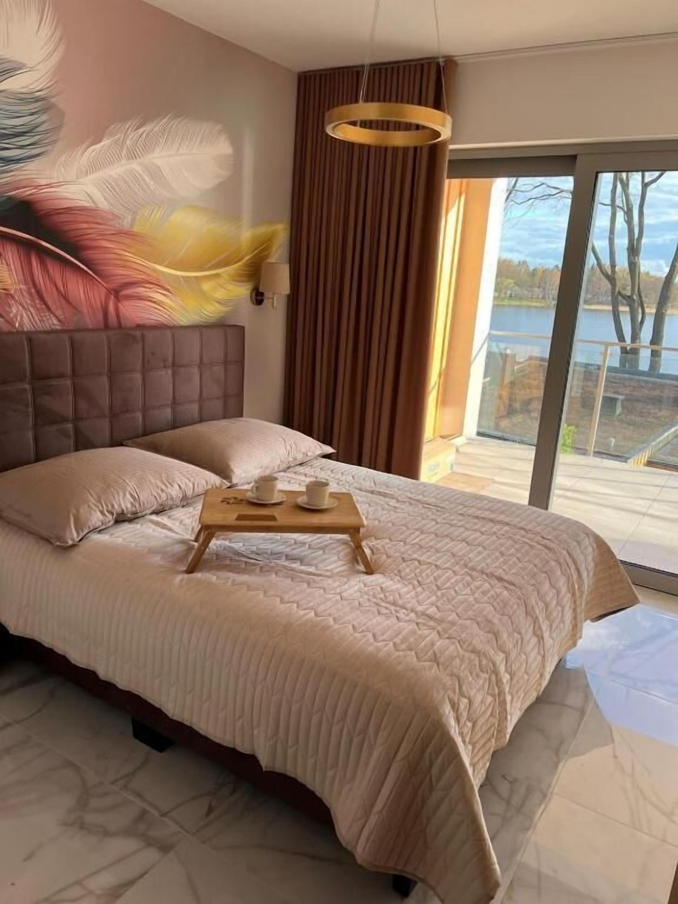 Ein Schlafzimmer mit einem großen Bett, einem Holztablett mit Tassen, einem Kopfteil, Wandmalerei und Blick auf einen See durch eine Glastür.