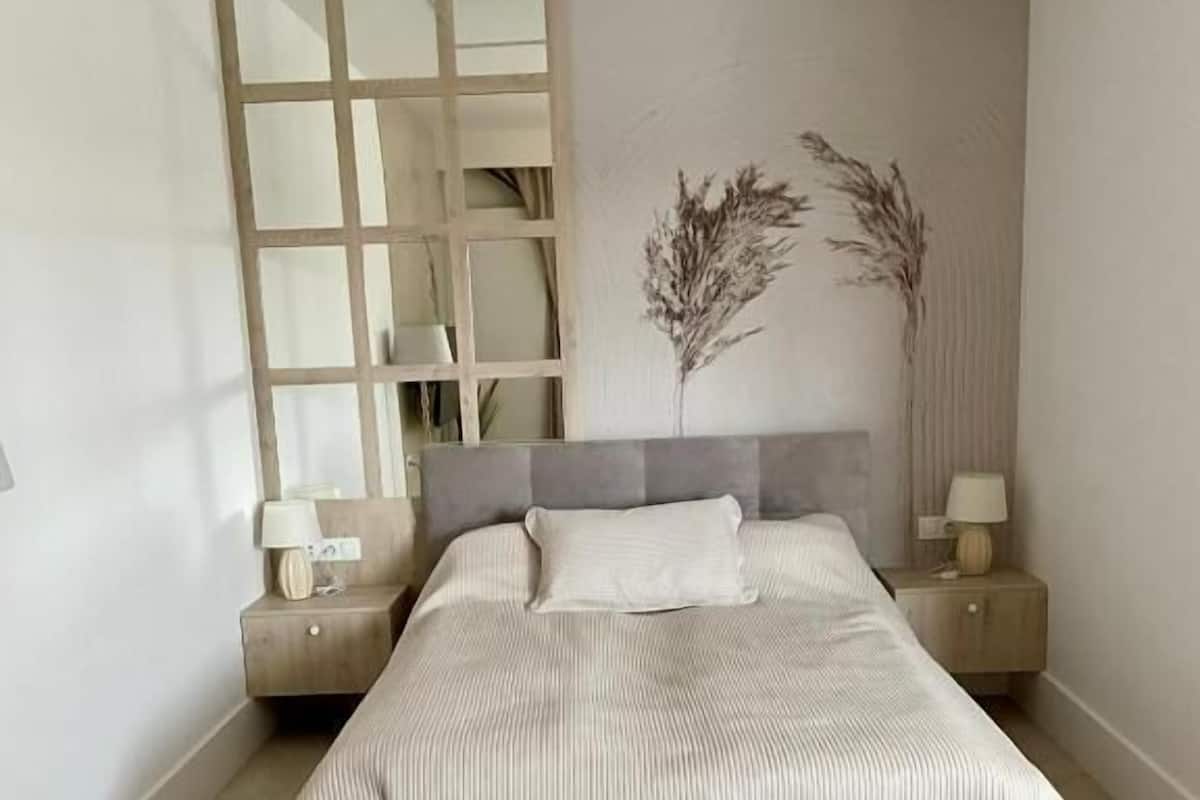 A bedroom with a bed, two bedside tables, a nightstand, a wall-mounted mirror, and a decorative wall piece.