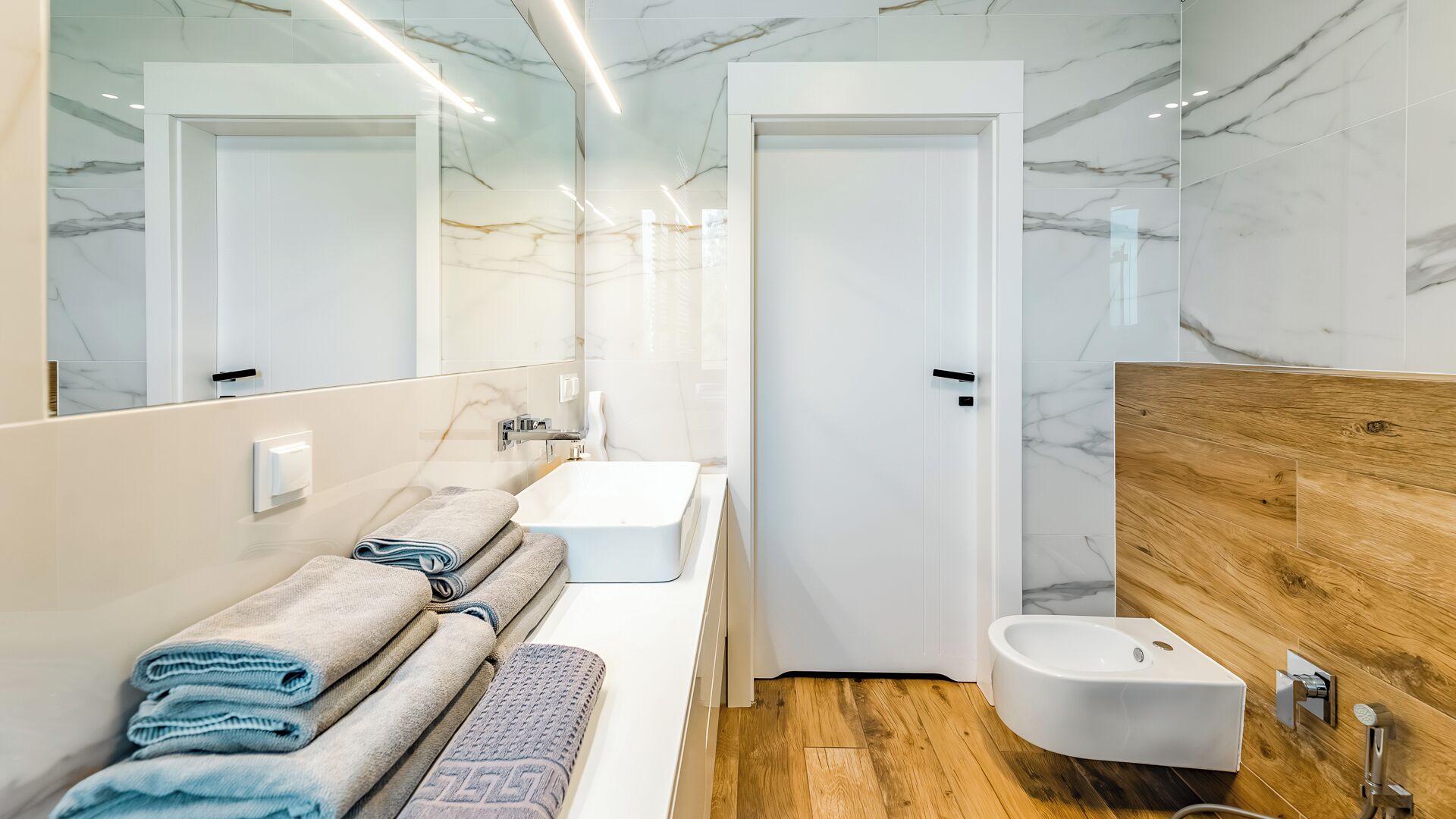 Un baño moderno con pared de mármol, tocador de madera, lavamanos blanco, bidé y espejo.