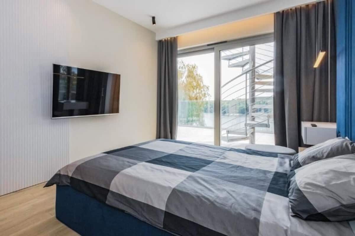 A modern bedroom with a large bed, a flat-screen TV mounted on the wall, and a balcony with a view of a building.