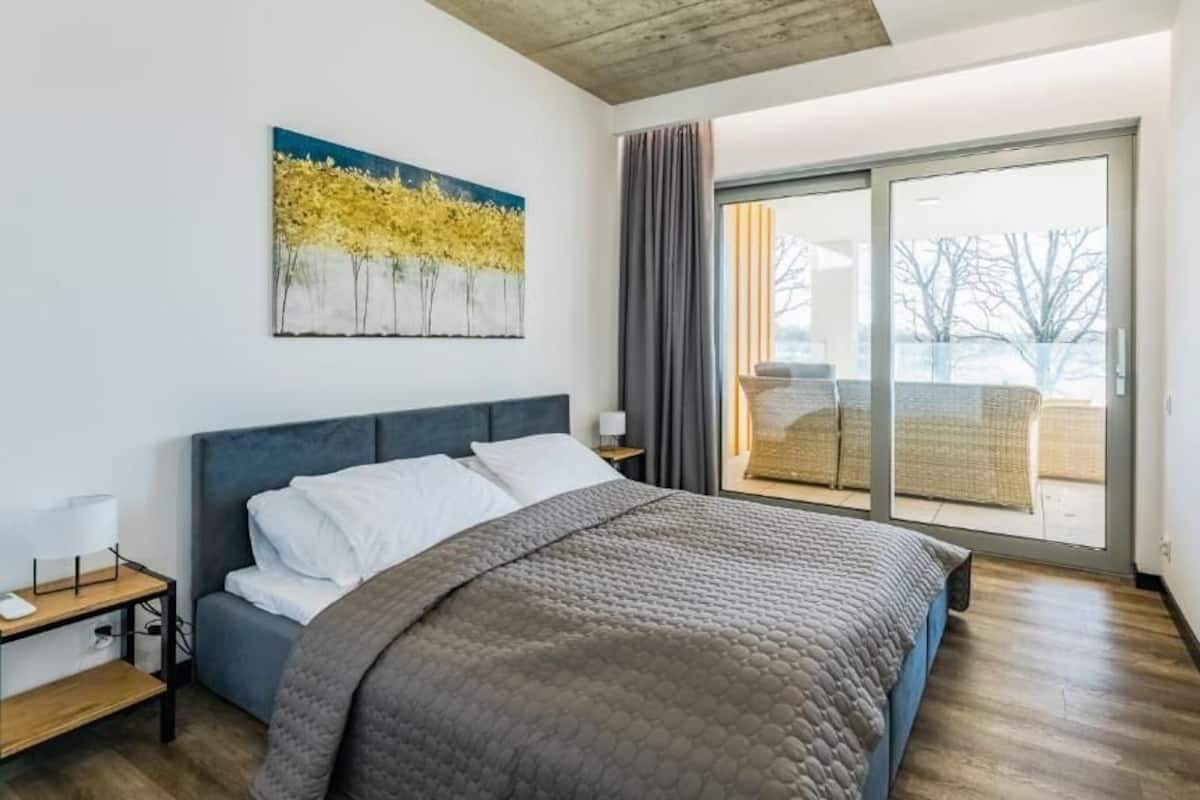 A bedroom with a bed, a nightstand, a painting, a sliding glass door to a balcony, and a wooden floor.