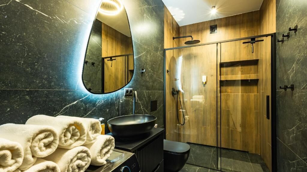Ein modernes Badezimmer mit großem Spiegel, einem schwarzen Waschbecken, einer Duschkabine aus Holz und ordentlich gerollten Handtüchern.
