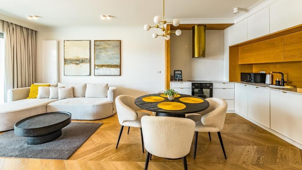 A modern living room with a sofa, dining table, and kitchen area with white cabinets and a wooden accent wall.