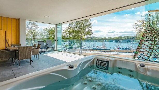 Un área interior de piscina moderna con vista a embarcaciones en el agua, un jacuzzi y una zona de estar con sillones.