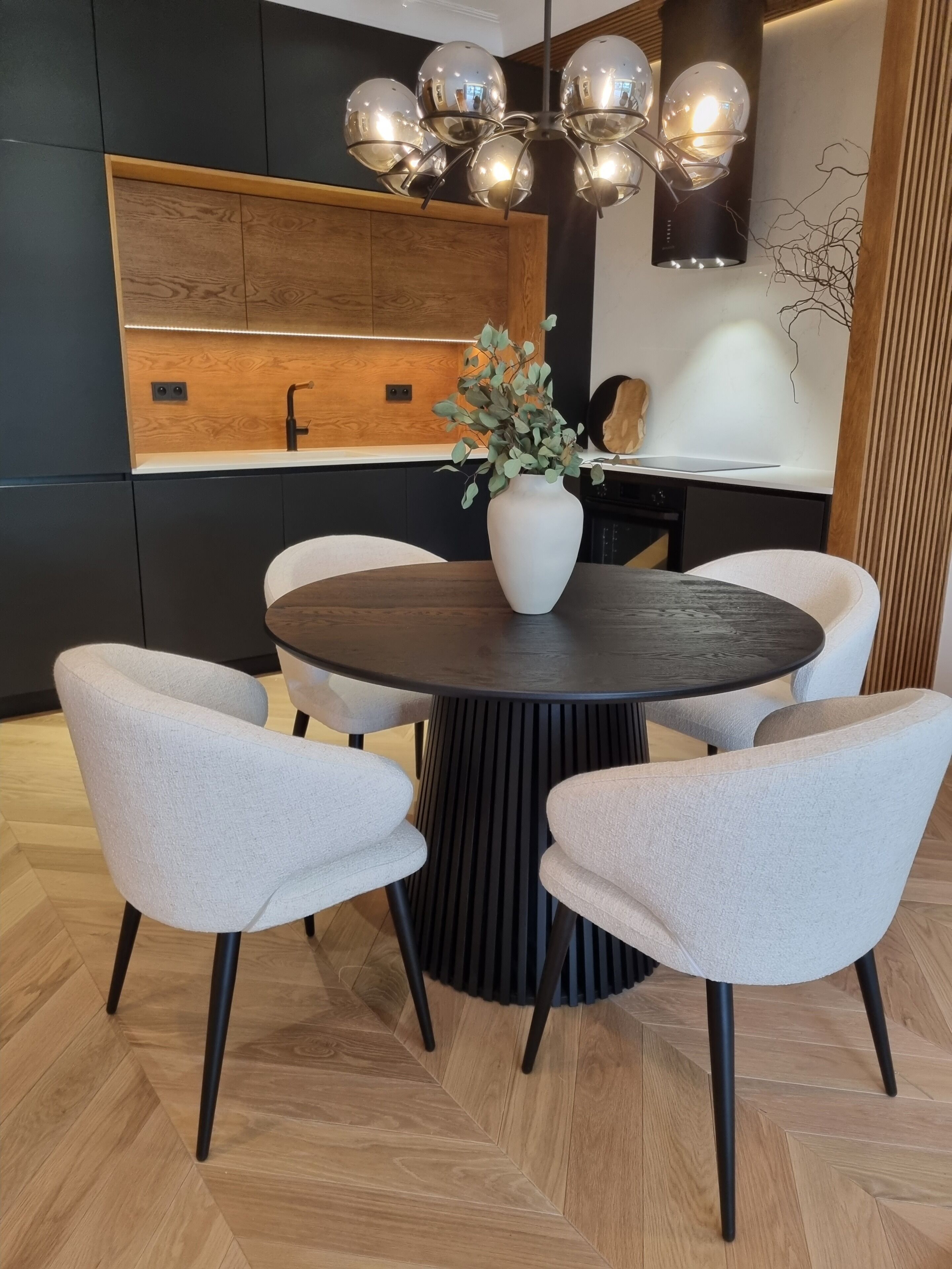 Un comedor moderno con una mesa redonda, cuatro sillas, un jarrón con follaje, una lámpara esférica original y una pared de acento de madera.