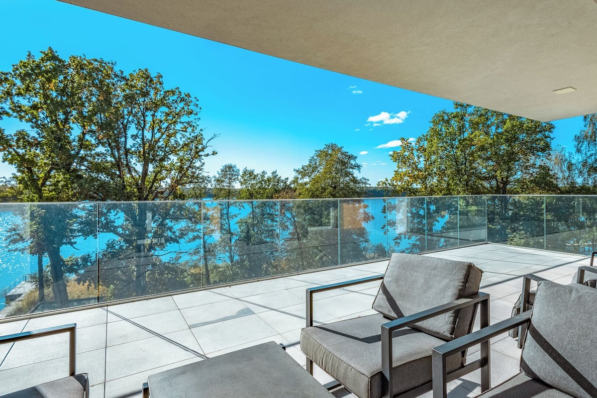 A modern outdoor seating area with a glass balustrade overlooking a body of water and trees.