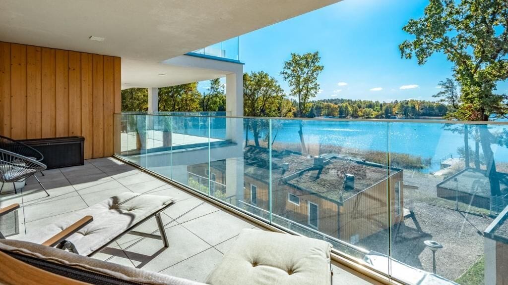 A balcony with a glass railing overlooking a lake, furnished with a sofa, chairs, and a table.