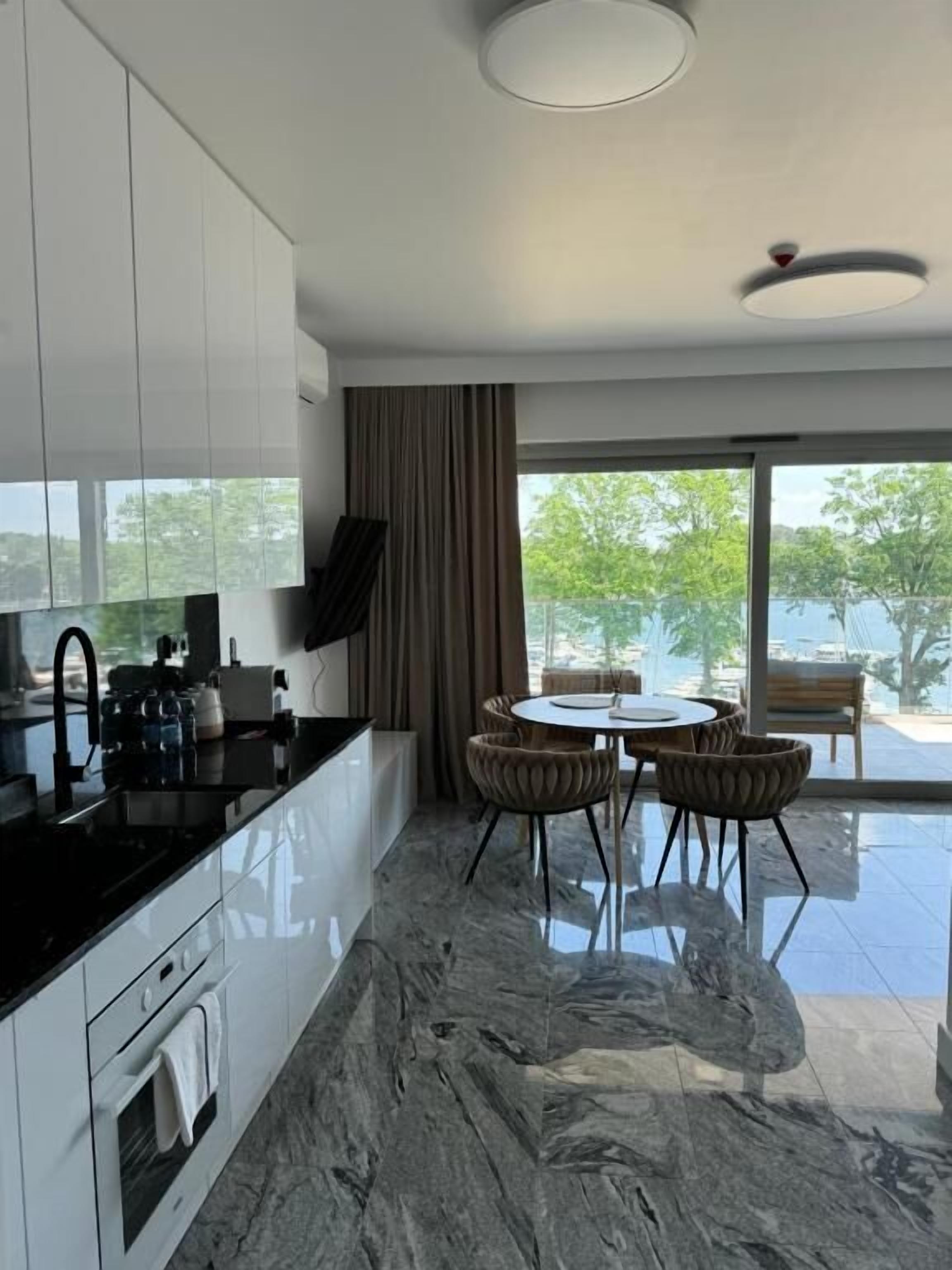 A modern kitchen with a marble floor, a dining area with a round table and chairs, and a large window with a view of greenery.