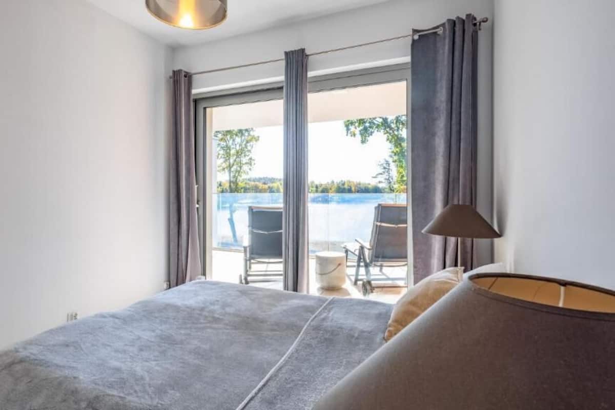 A bedroom with a bed, a chair, a table, and a view of a balcony with a chair and a table.