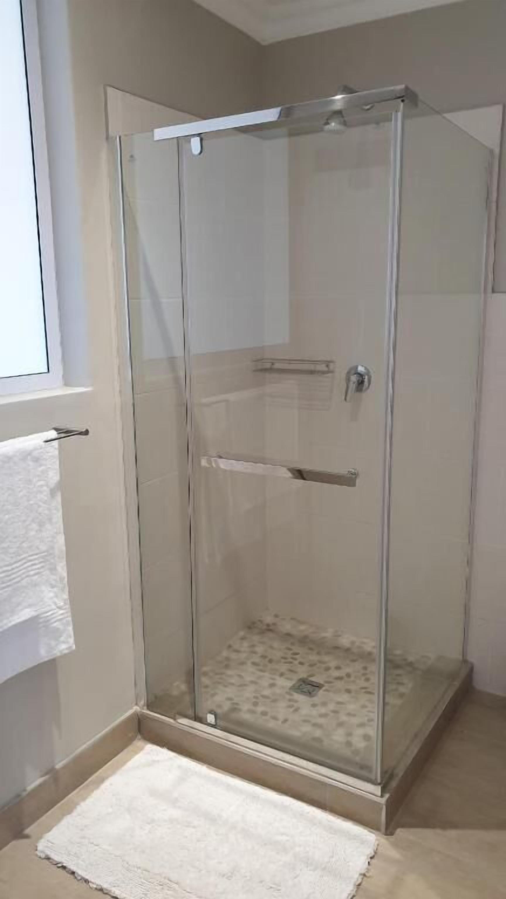 A glass-enclosed shower with a white towel hanging on the door, a small shelf inside, and a white rug on the floor.