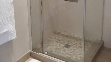 A glass-enclosed shower with a white towel hanging on the door, a small shelf inside, and a white rug on the floor.