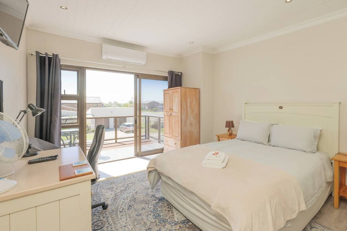A hotel room with a bed, bedside tables, a desk with a chair, a fan, and a view of the outside.