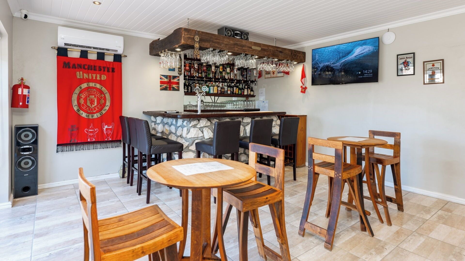 Zona de bar con pared de piedra, sillas de madera, barra y una pantalla grande.