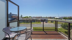 Un balcon avec une table et des chaises en verre, une vue sur une rue résidentielle et un ciel dégagé.