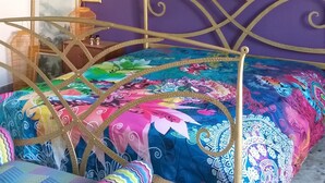 Un dormitorio con pared morada, cama con colcha colorida, candelabro y cuadros enmarcados.